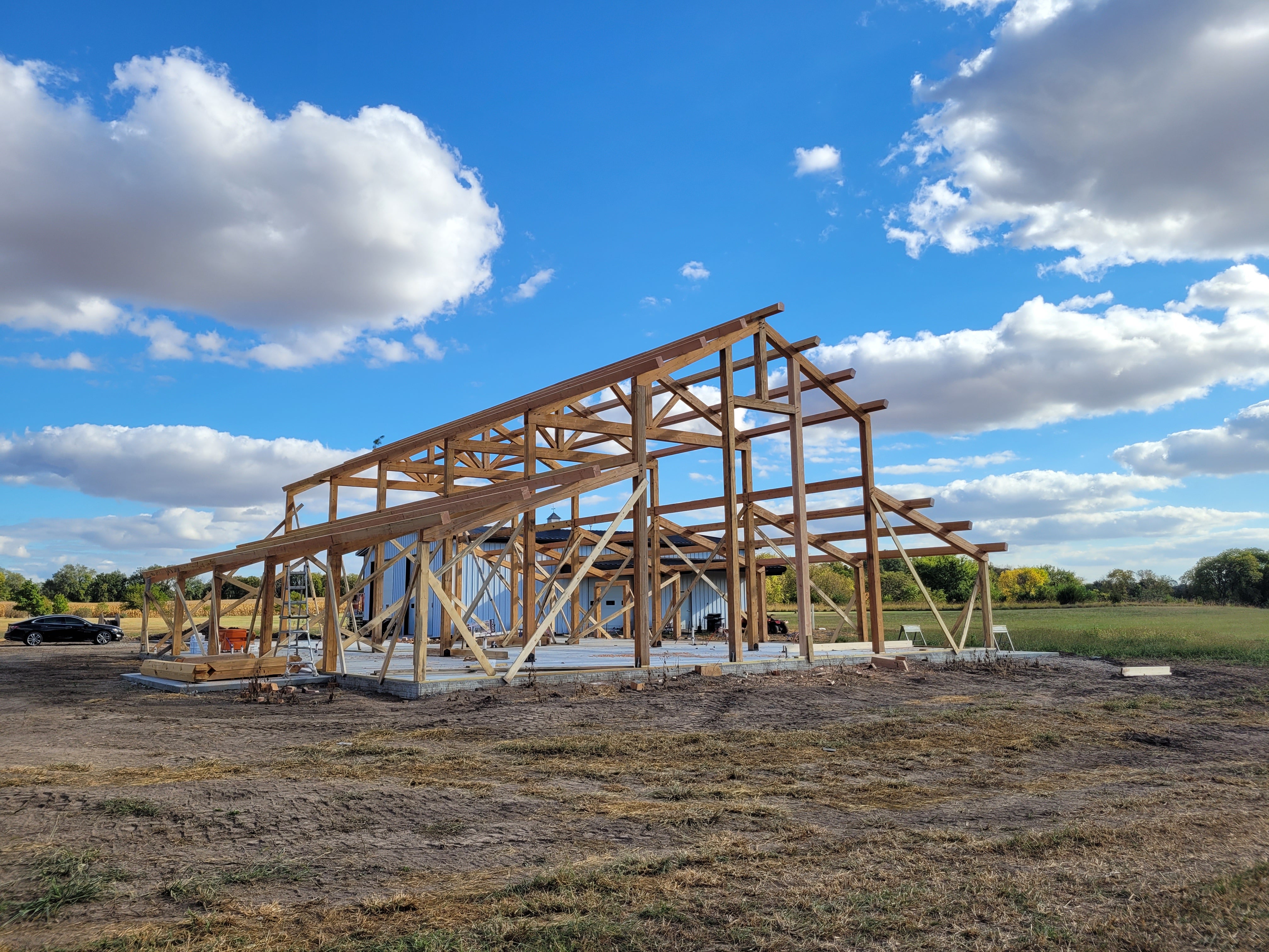 A large wooden barn or shed frame under construction, with exposed timber beams and a roof structure, set in an open field under a blue sky with white clouds.