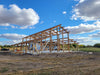 A large wooden barn or shed frame under construction, with exposed timber beams and a roof structure, set in an open field under a blue sky with white clouds.