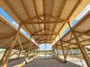 A large open wooden structure under construction, viewed from inside looking up at a steep gable roof with exposed wooden beams and rafters against a blue sky.