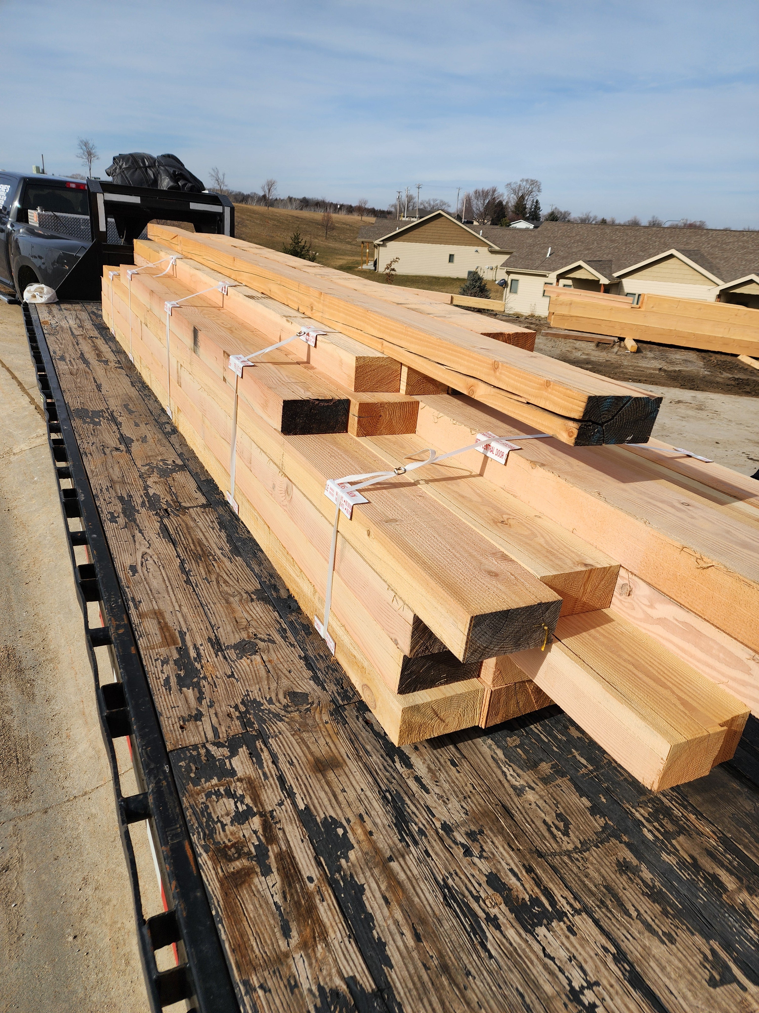 Large stacks of raw lumber and plywood sheets secured on a flatbed truck in an outdoor lot, with houses and a blue sky visible in the background.