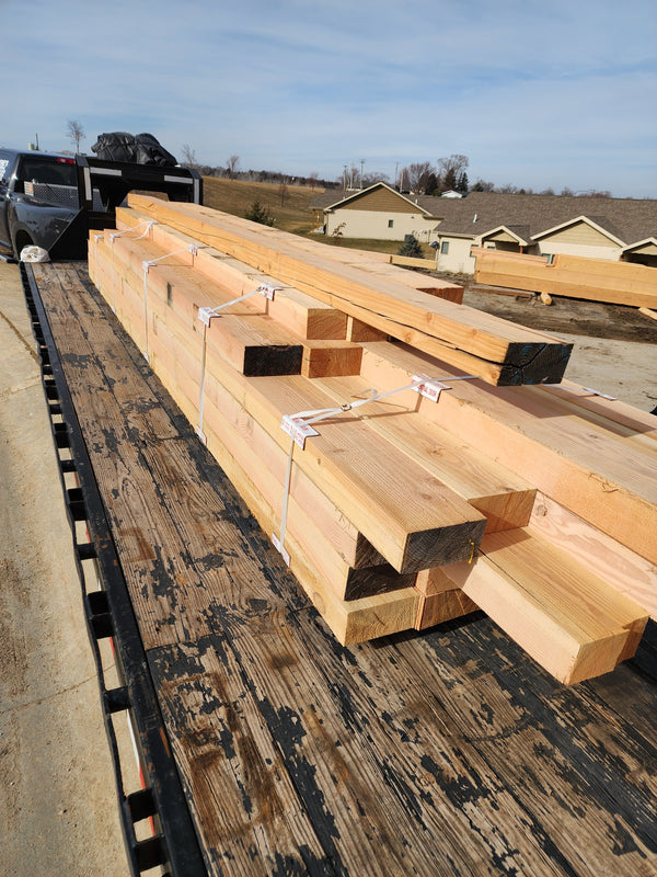 Large stacks of raw lumber and plywood sheets secured on a flatbed truck in an outdoor lot, with houses and a blue sky visible in the background.