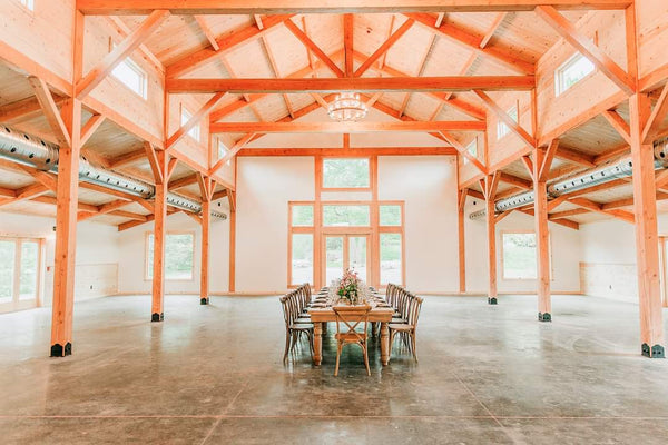 A large, empty barn interior with tall orange-painted wooden beams and columns, a concrete floor, and a long dining table with chairs centered near the front windows.