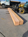 Two large natural wood beams stacked on a concrete surface in an outdoor industrial yard, with a yellow forklift and a dump truck in the background.
