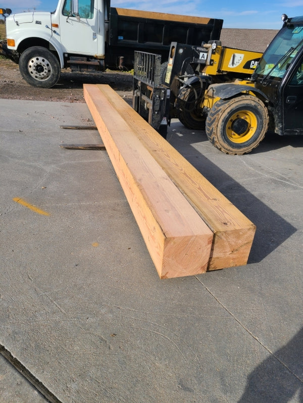 Two large natural wood beams stacked on a concrete surface in an outdoor industrial yard, with a yellow forklift and a dump truck in the background.